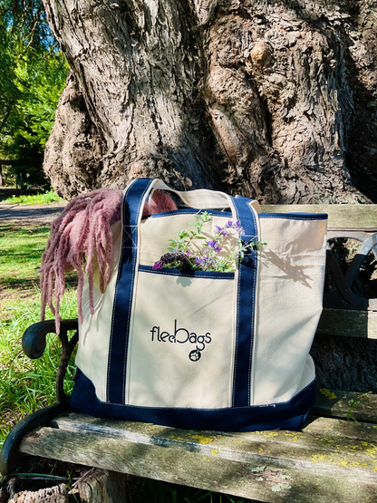 Beige tote bag with navy blue trimming and 'Fleebags' branding on a wooden bench outdoors.